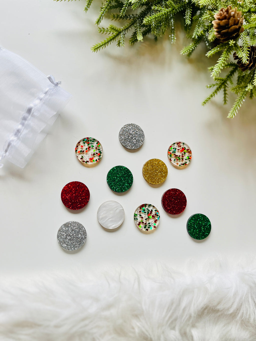 Collection of round acrylic shapes in glittery red, green, gold, silver, pearlescent white, and confetti patterns on white surface with greenery