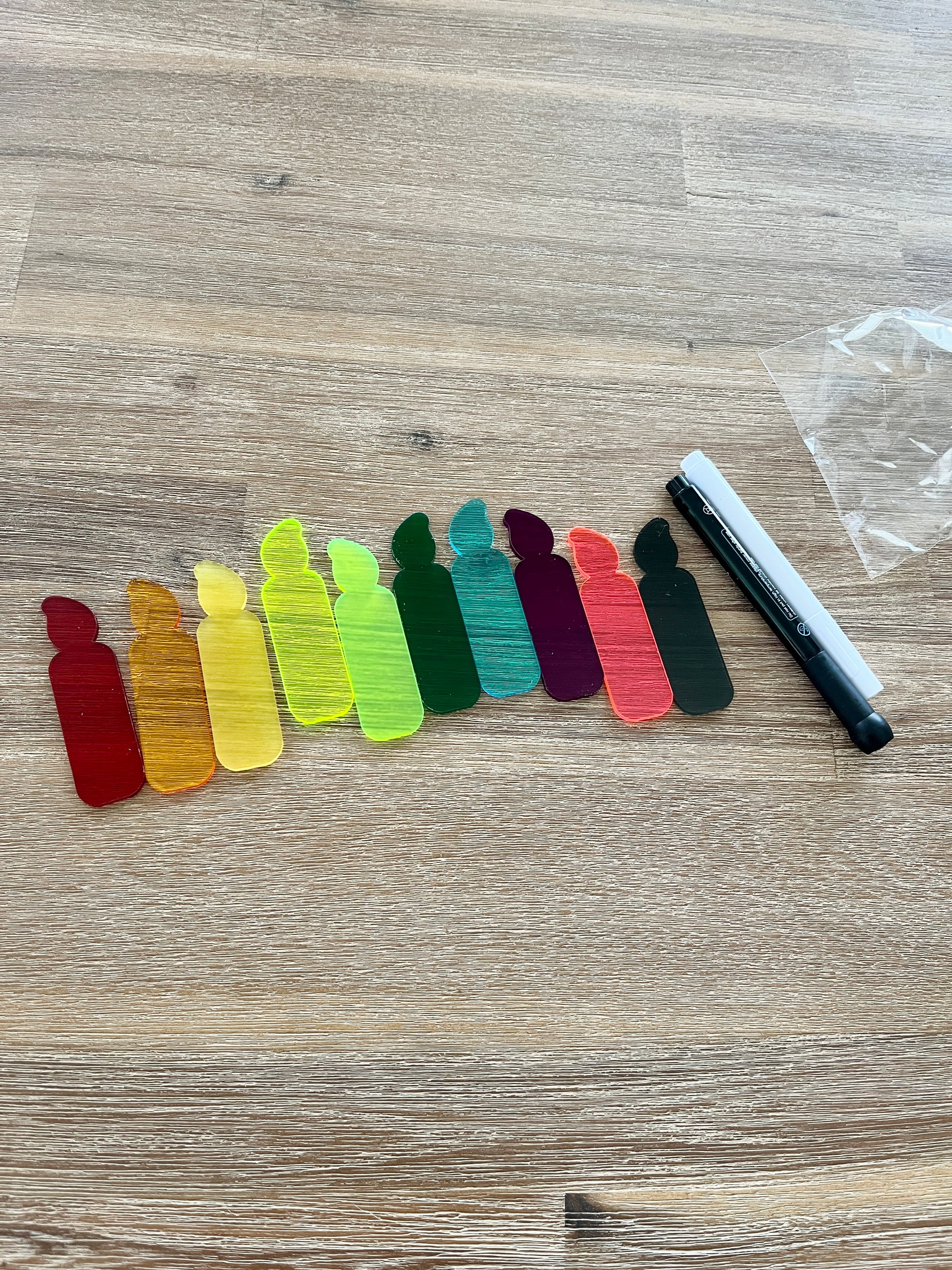 A row of colorful translucent candle-shaped pieces arranged on a wooden surface next to black and white dry erase markers, featuring Little Dry Erasables - Translucent Candles - Set of 5 or 10, handcrafted homeschool education resources.