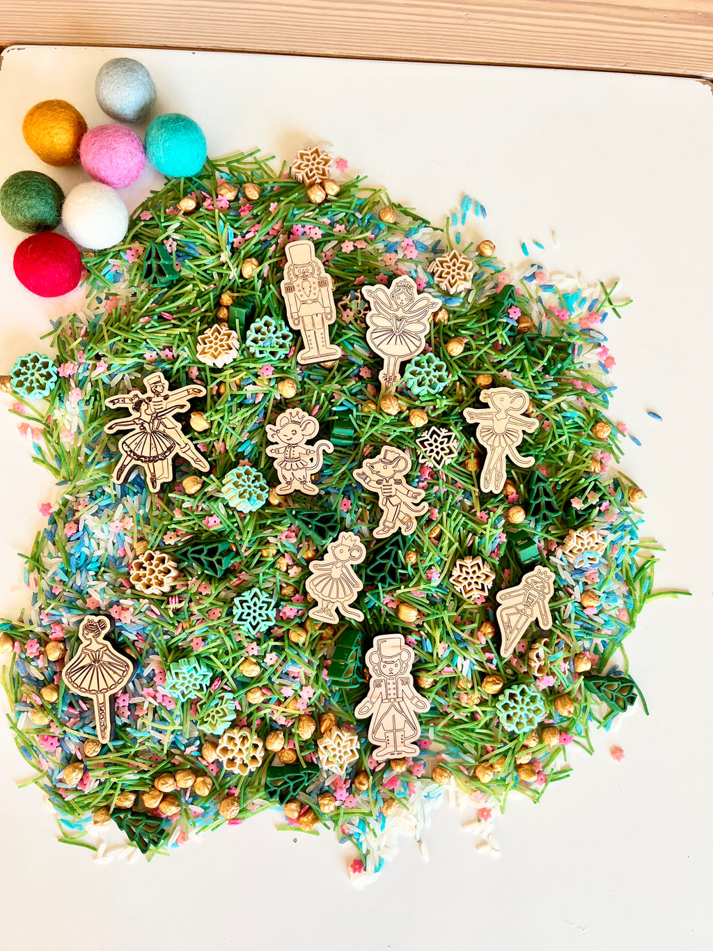 Wooden nutcracker and ballerina figurines on green pine needle mix with pastel sprinkles and gold beads on white surface