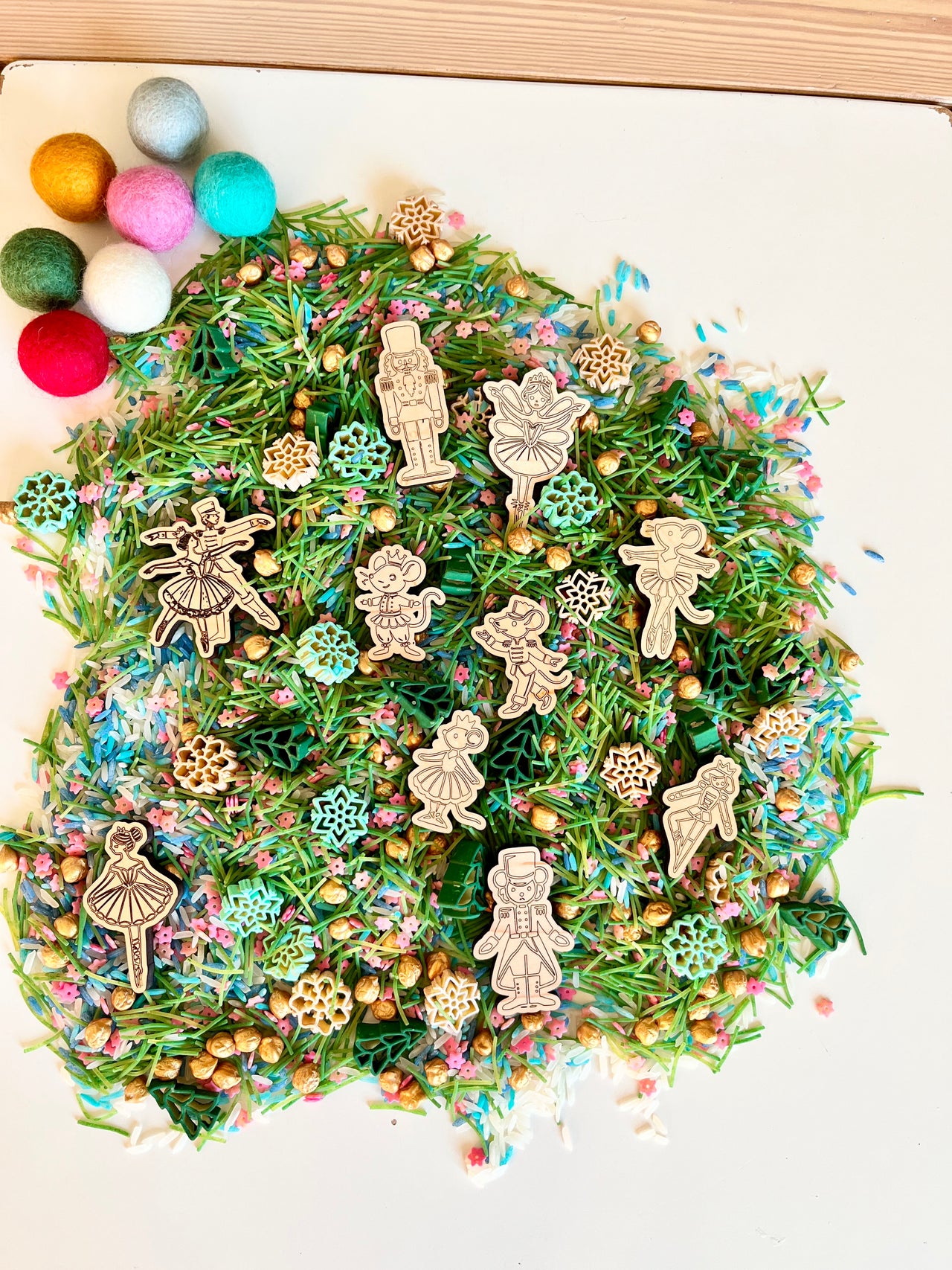 Wooden nutcracker and ballerina figurines on green pine needle mix with pastel sprinkles and gold beads on white surface