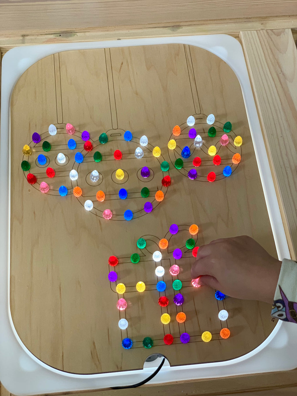 Wooden Flisat insert with Christmas ball and gift shapes lit up by colorful light pegs with child’s hand placing a peg
