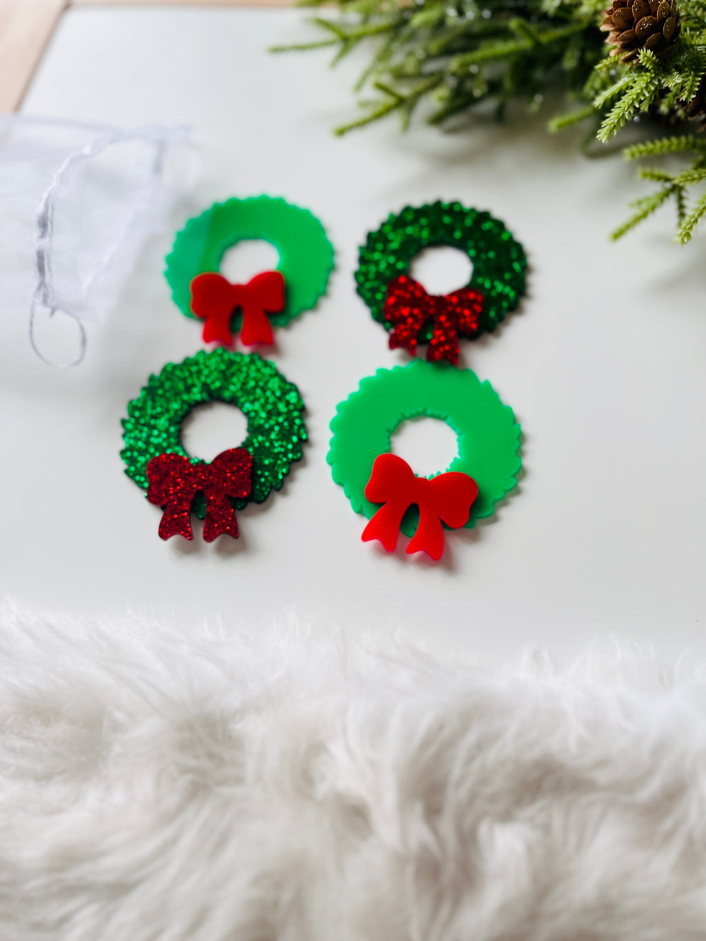 Four small green acrylic Christmas wreaths with red bows, two with solid finish and two glittery, on white surface with greenery nearby