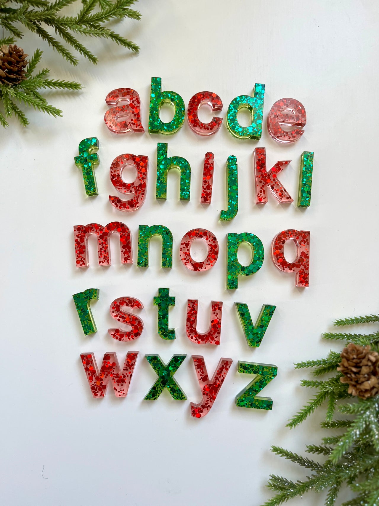 Red and green glitter-filled resin lowercase alphabet letters arranged on white surface with pine branches and cones nearby