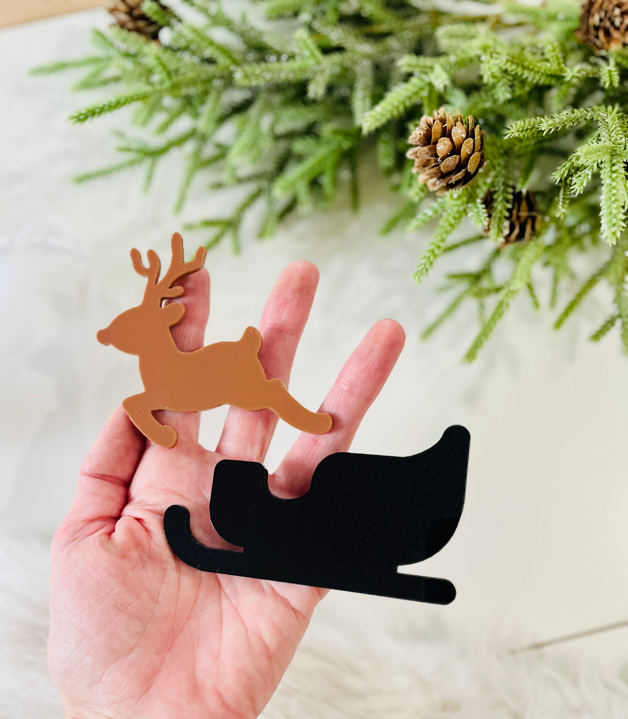 Hand holding flat brown reindeer and black sleigh-shaped acrylic decorations with pine branches and cones nearby