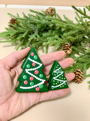Hand holding two small wooden Christmas trees painted with green, red, and white details with pine cones and branches background