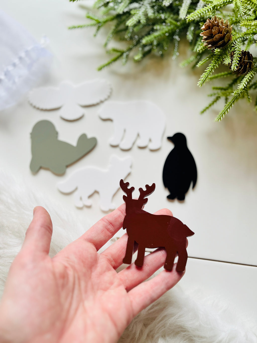 Hand holding brown moose-shaped acrylic cutout with flat animal shapes and greenery background on light surface