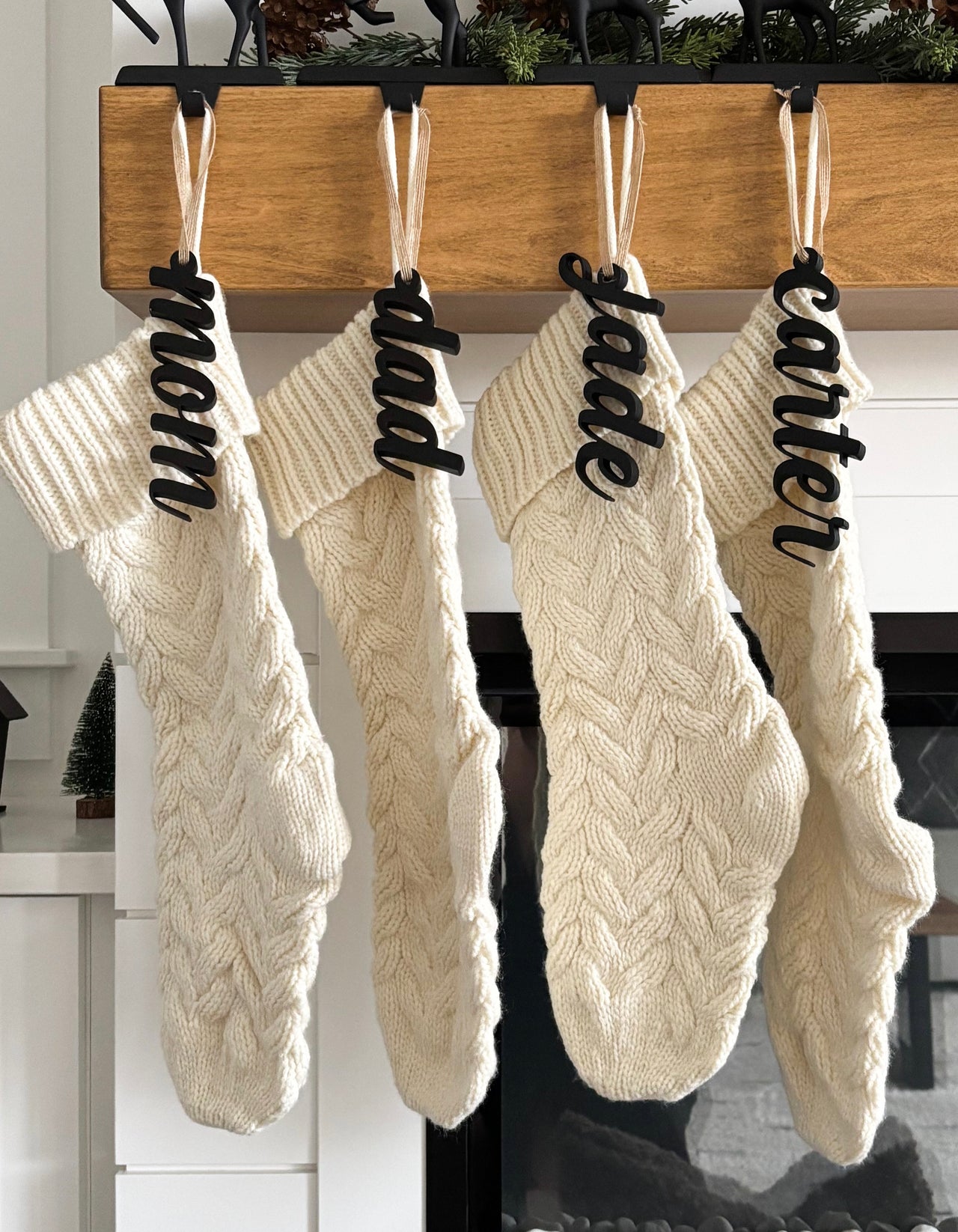 Four cream-colored cable knit Christmas stockings hanging from a wooden mantel with black name tags and white ribbons