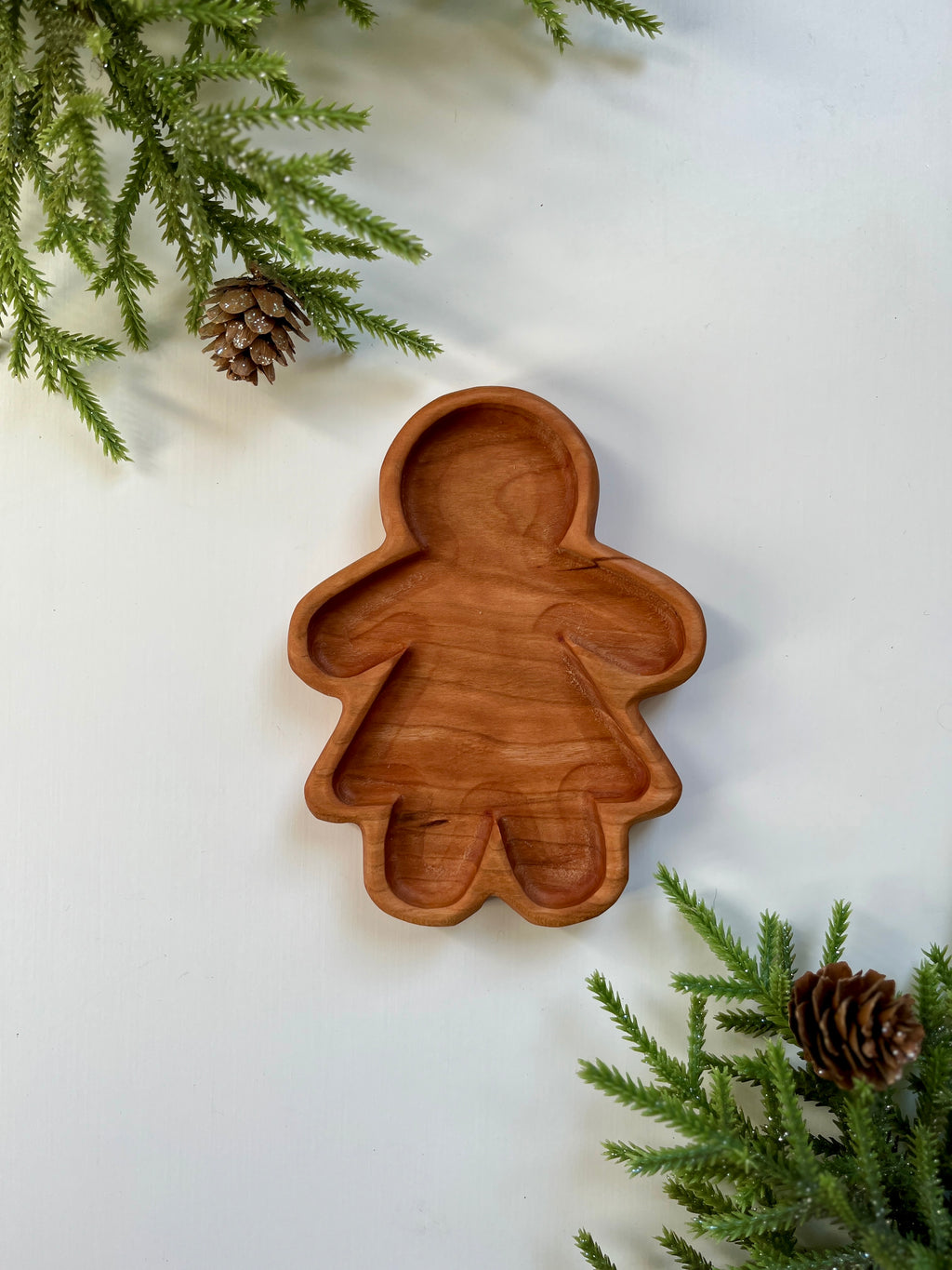 Carved wooden tray shaped like a gingerbread girl surrounded by pine branches and pine cones on white surface