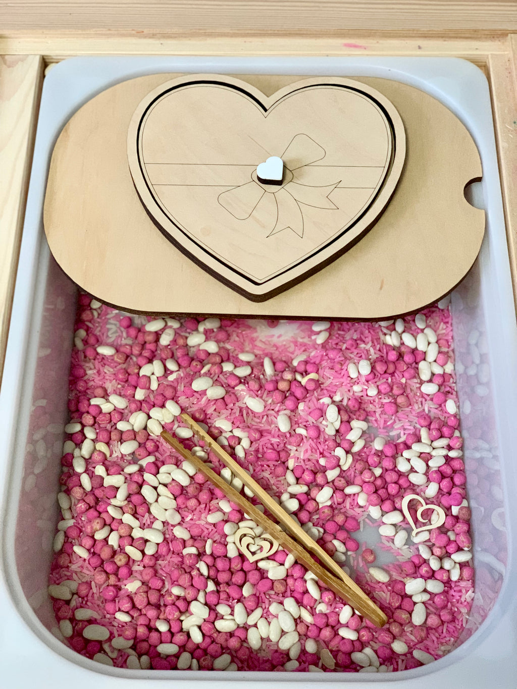 Plastic bin filled with pink rice, white beans, small wooden heart shapes, and wooden tweezers with heart-shaped lid above
