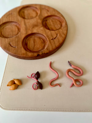 Round wooden tray with five circular indentations beside small realistic plastic worms and seeds on a cream surface
