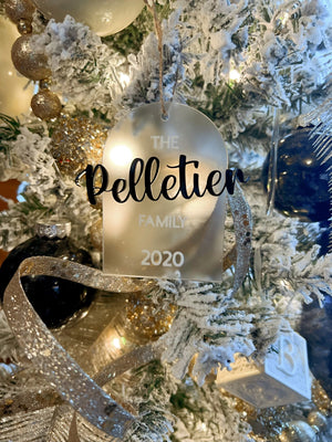 Frosted acrylic arch ornament with black and white text displaying "The Pelletier Family 2020" hanging on snowy branches