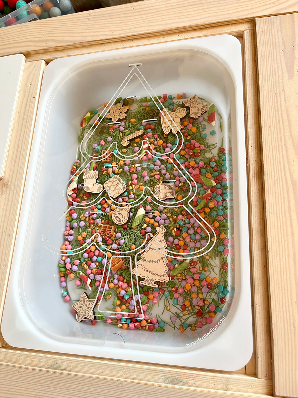 Christmas tree-shaped tray filled with colorful small beads, dried greenery, and wooden holiday-themed cutouts on wooden surface
