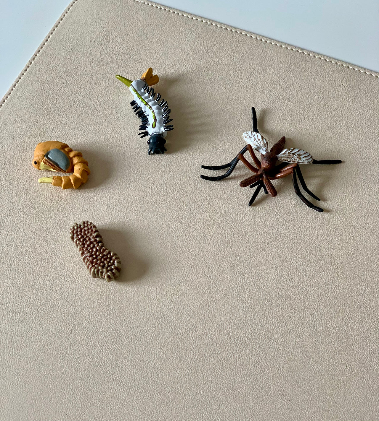 Four detailed insect and larva figurines including a mosquito, pupa, and segmented caterpillar on beige surface