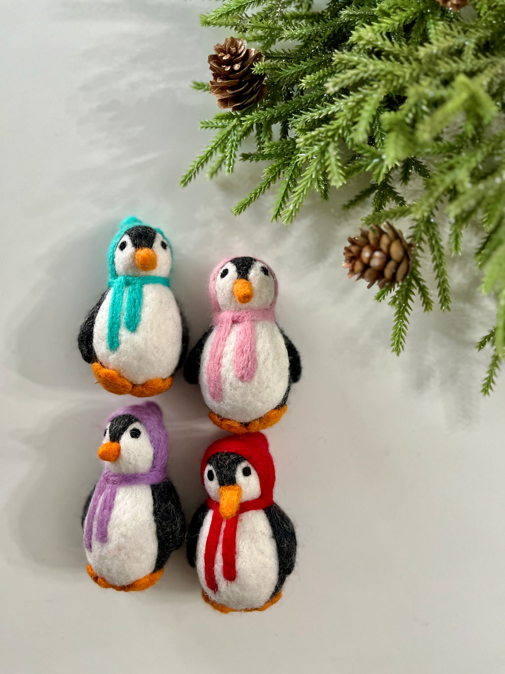 Four felt penguin ornaments wearing colorful scarves arranged near pine branches with cones on a white background