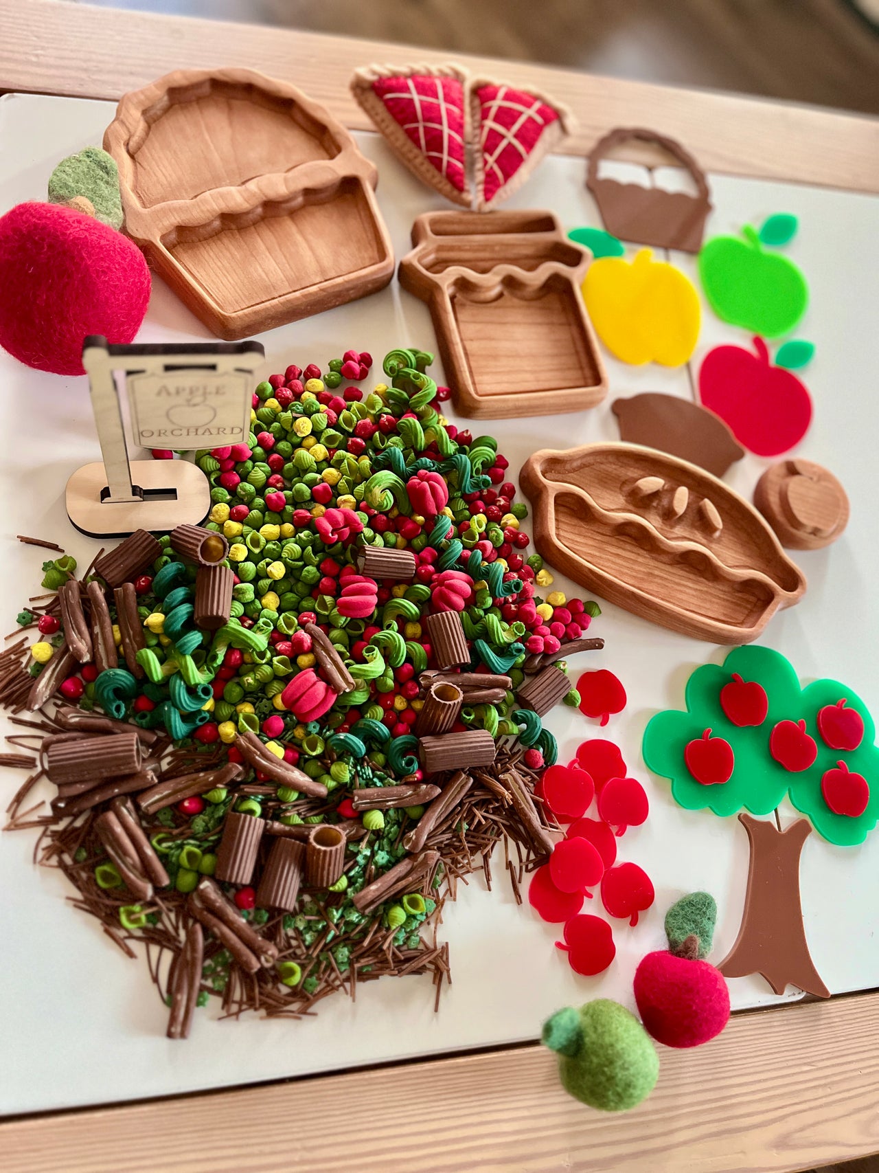 Miniature clay pasta and felt apples arranged beside wooden trays and felt pie slices on a white surface
