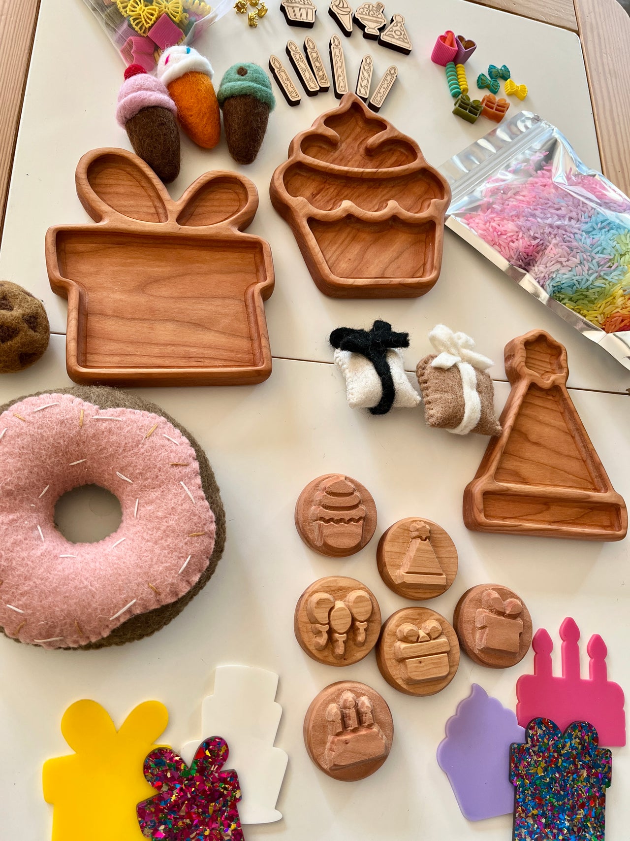 Wooden trays shaped like a gift box, cupcake, and party hat with felt desserts, colorful sprinkles, and craft stamps on table