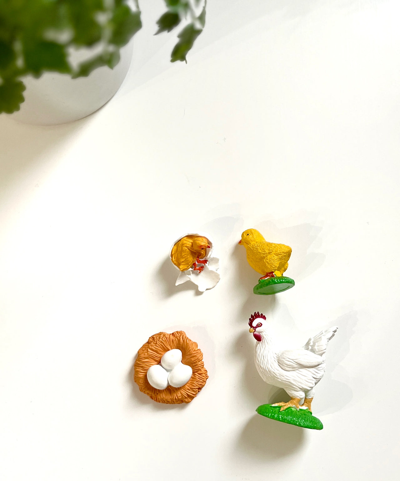 Small figurines of a white hen, yellow chicks, and a nest with white eggs on a white surface near green plant leaves