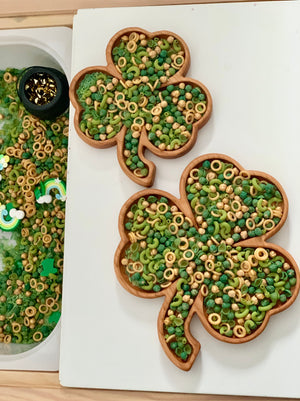 Two wooden shamrock-shaped trays filled with green and beige pasta, beads, and small decorative pieces on white surface