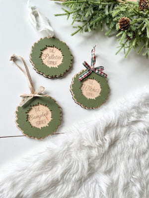 Three personalized wooden wreath ornaments with green leaves, scalloped edges, and plaid or ribbon bows on white surface