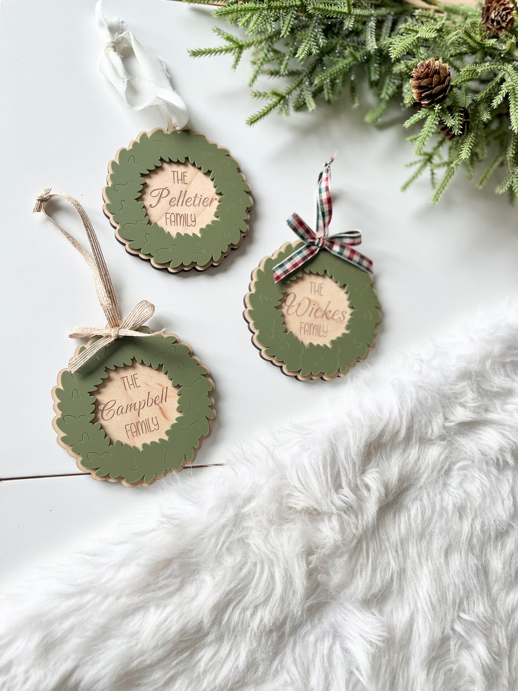 Three personalized wooden wreath ornaments with green leaves, scalloped edges, and plaid or ribbon bows on white surface