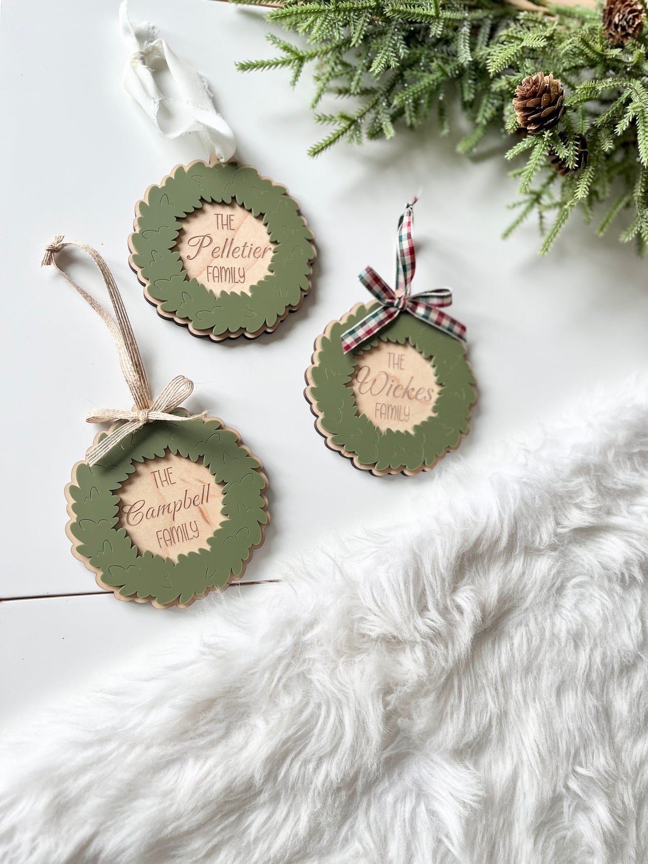 Three personalized wooden wreath ornaments with green leaves, scalloped edges, and plaid or ribbon bows on white surface