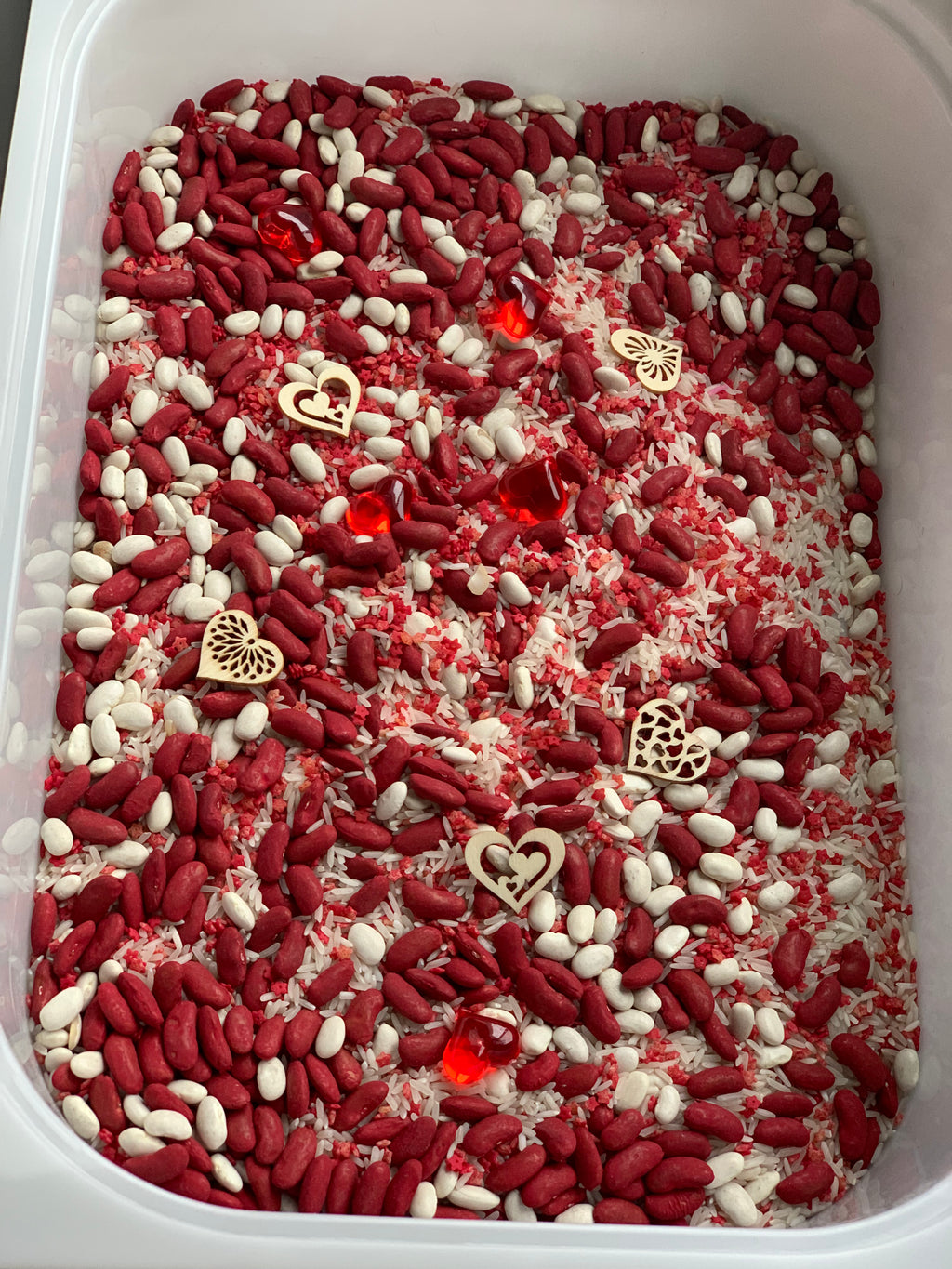 White and red beans mixed with dyed rice and scattered wooden and plastic heart-shaped decorations in a white container