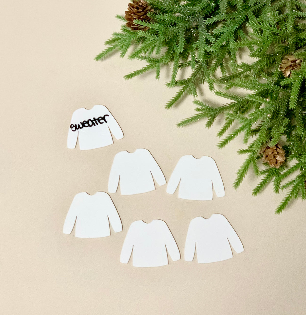 Set of six white sweater-shaped dry erase boards with one written "sweater" beside pine branches and cones on beige surface