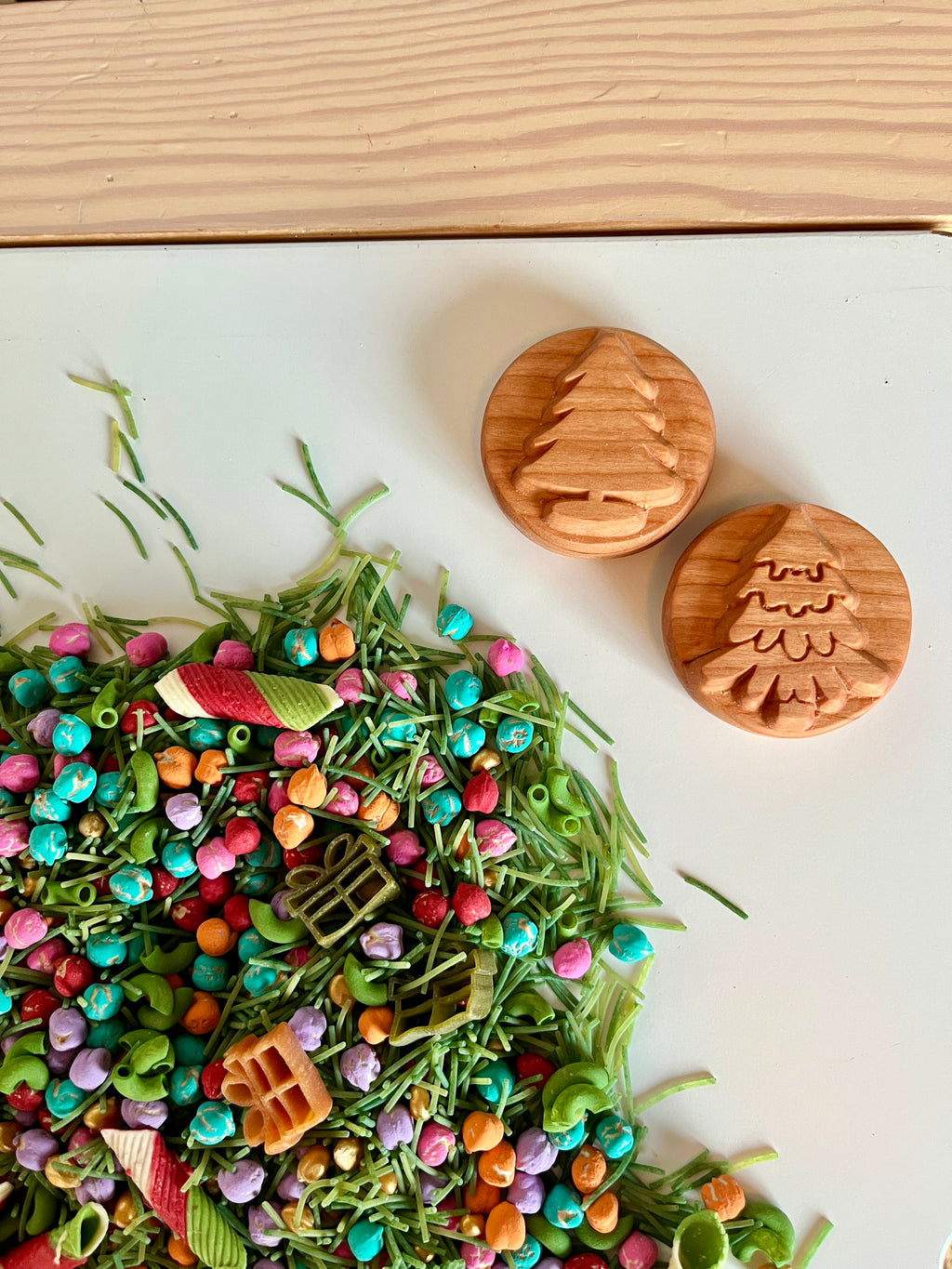 Two wooden circular stamps with embossed Christmas tree designs beside colorful pasta and green straw bits on white surface