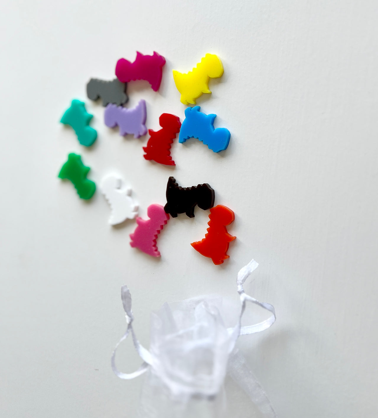 Assorted colorful plastic dinosaur-shaped game pieces scattered above a small sheer white drawstring pouch on a white background