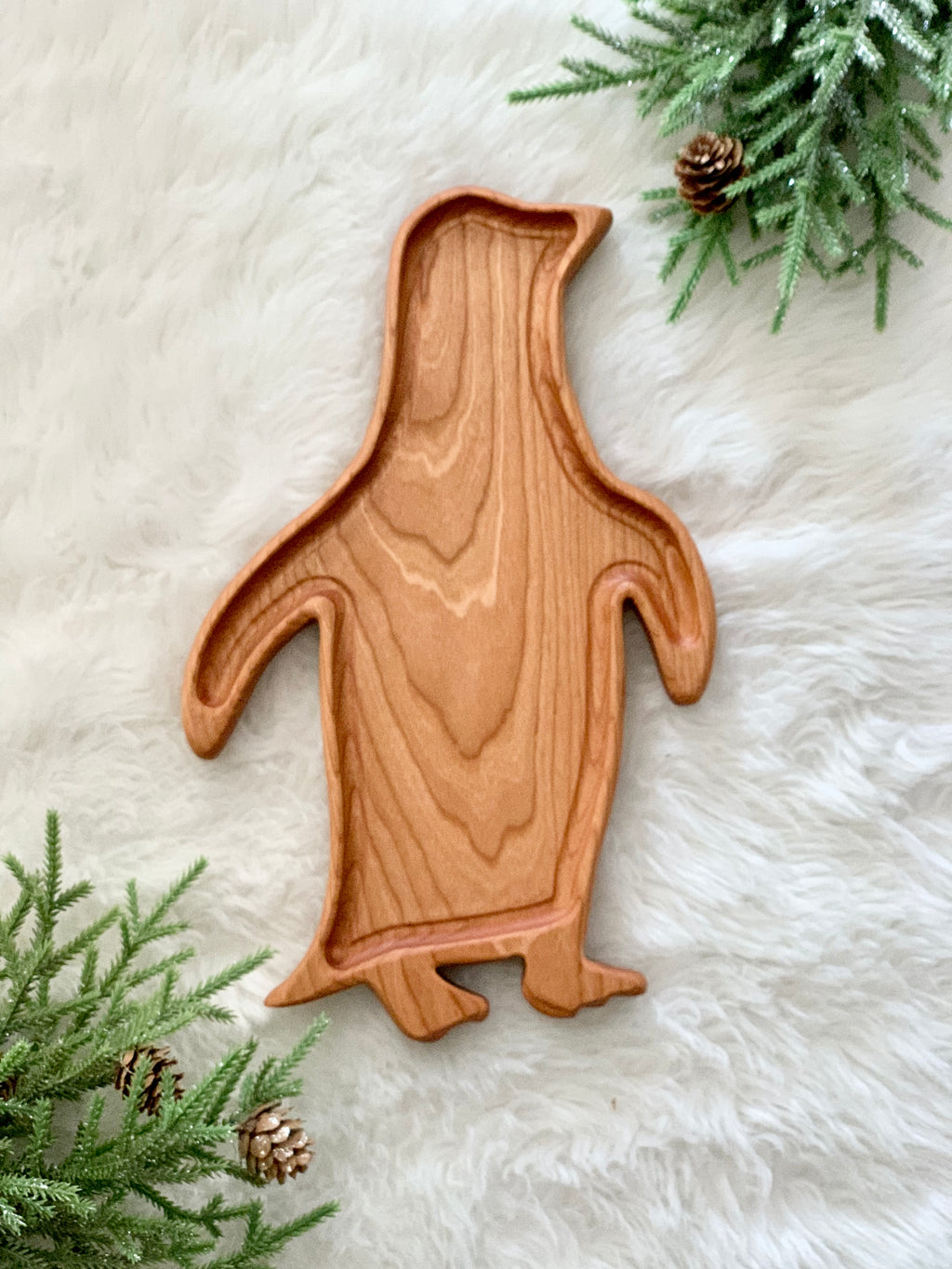 Penguin-shaped wooden sensory tray on white faux fur with green pine branches and pinecones nearby