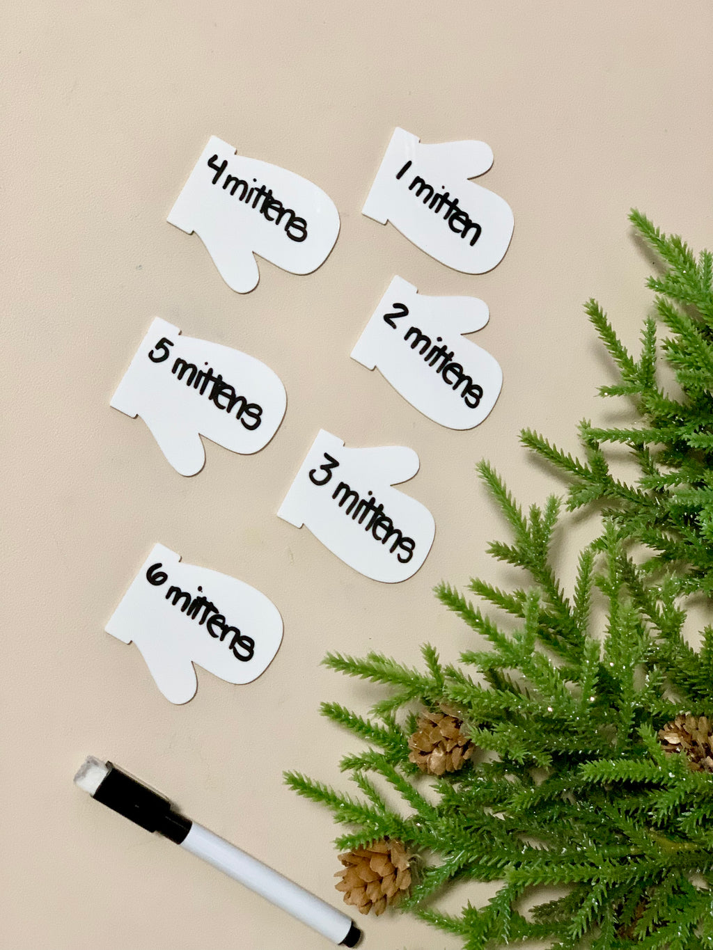 Set of six white mitten-shaped dry erase cards numbered one to six with black marker pen and pine branches on beige surface
