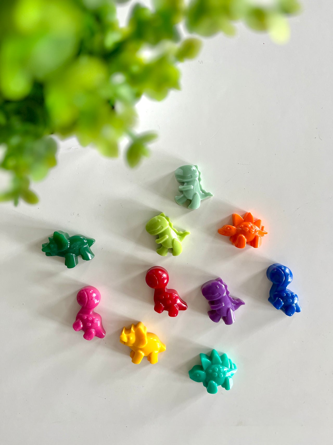 Nine colorful dinosaur-shaped plastic figures arranged on a white surface with blurred green leaves in the background
