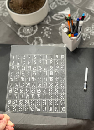 Hand holding transparent acrylic sheet with numbered grid 1 to 100 over a dark table with markers and plant nearby
