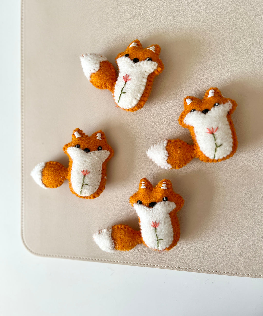 Four small felt fox toys with embroidered flowers on cream and orange fabric on beige surface