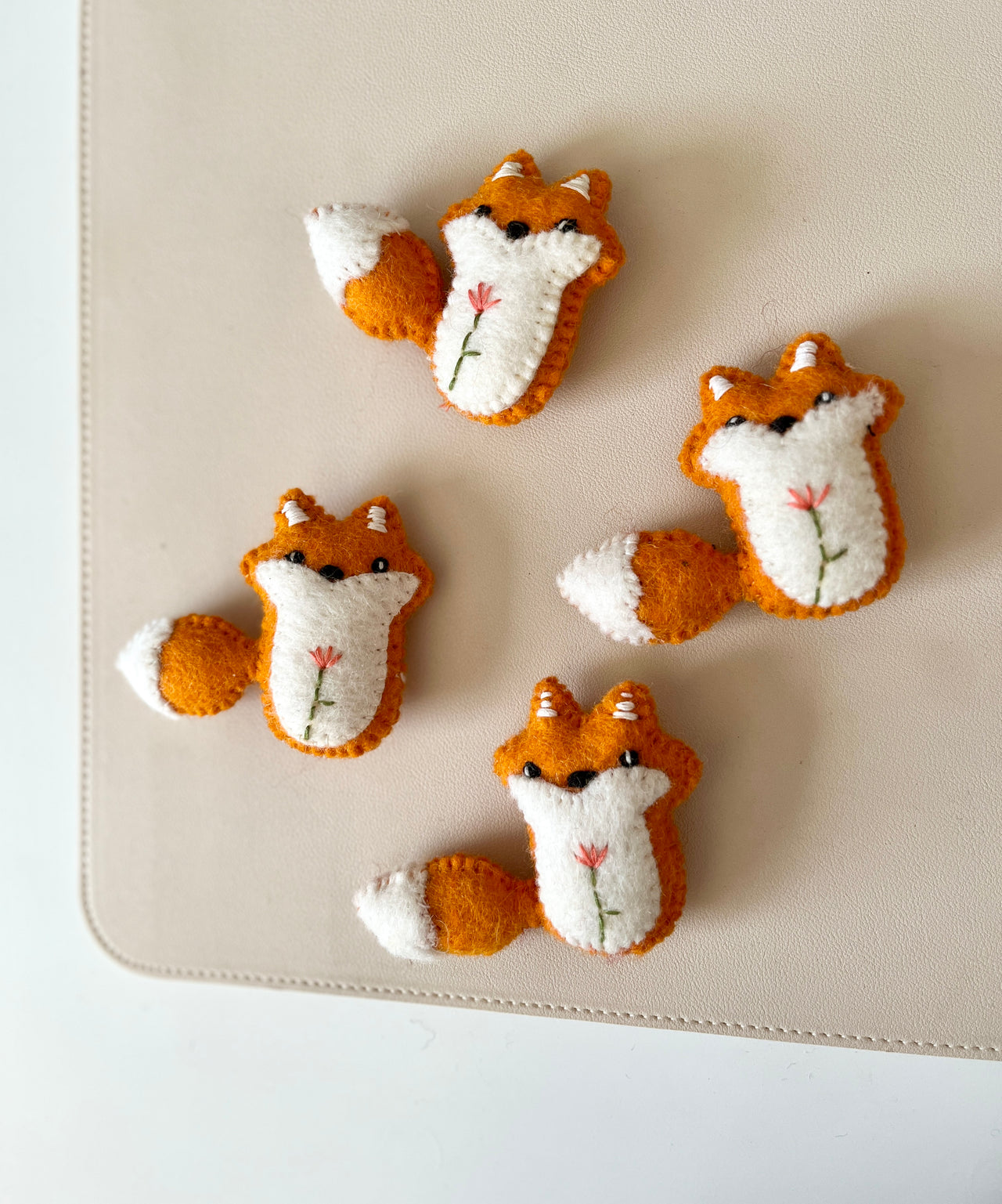 Four small felt fox toys with embroidered flowers on cream and orange fabric on beige surface