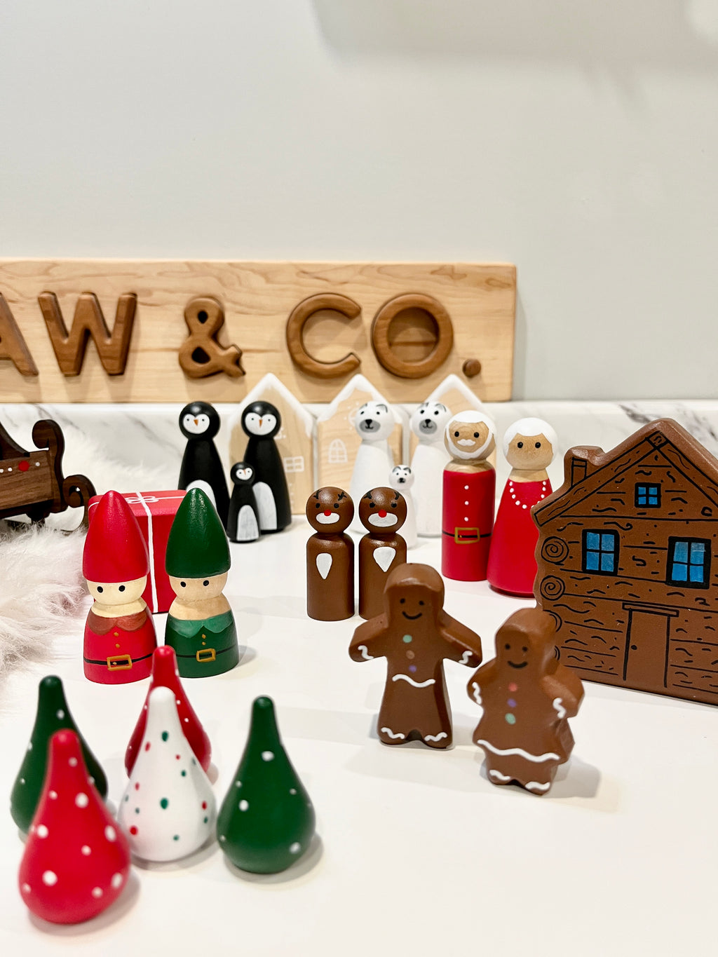Wooden holiday figures including elves, gingerbread people, polar bears, penguins, and a carved log cabin on white surface