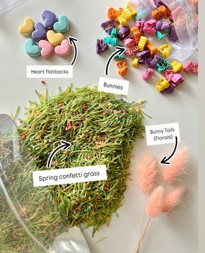 Pastel-colored heart-shaped pieces, colorful bunny figurines, pink bunny tail florals, and green confetti grass scattered on white surface