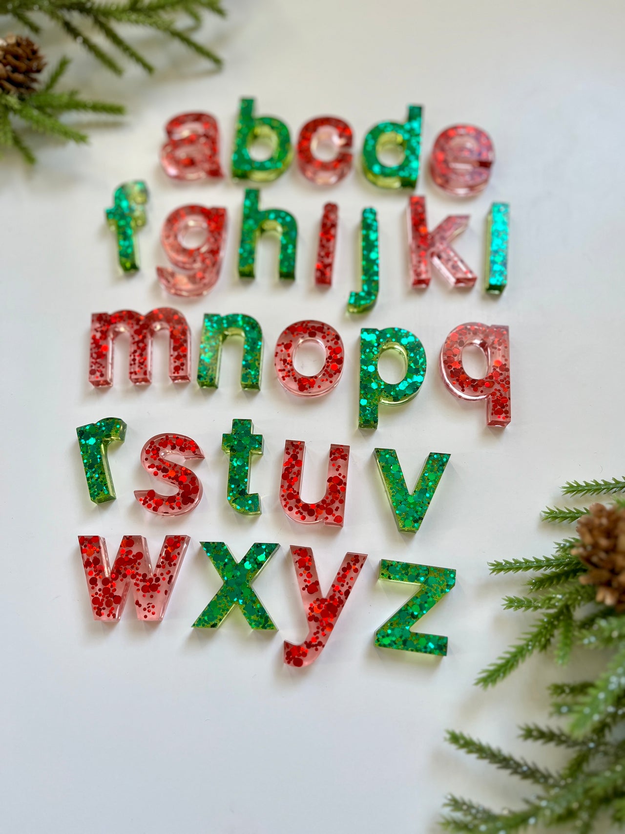 Red and green glitter-filled lowercase resin alphabet letters on white surface with pine branches and pine cones around edges