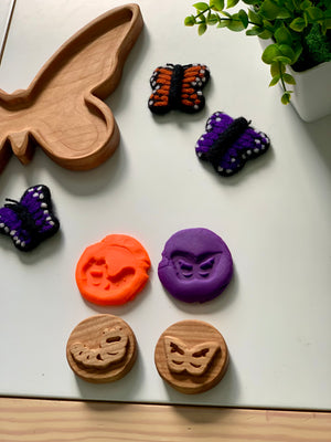 Wooden butterfly tray with felted butterfly toys, butterfly-shaped clay impressions, and wooden stamps on white surface with plant nearby