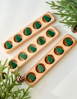 Three wooden mancala boards with green Christmas tree-shaped playing pieces surrounded by pine branches and cones