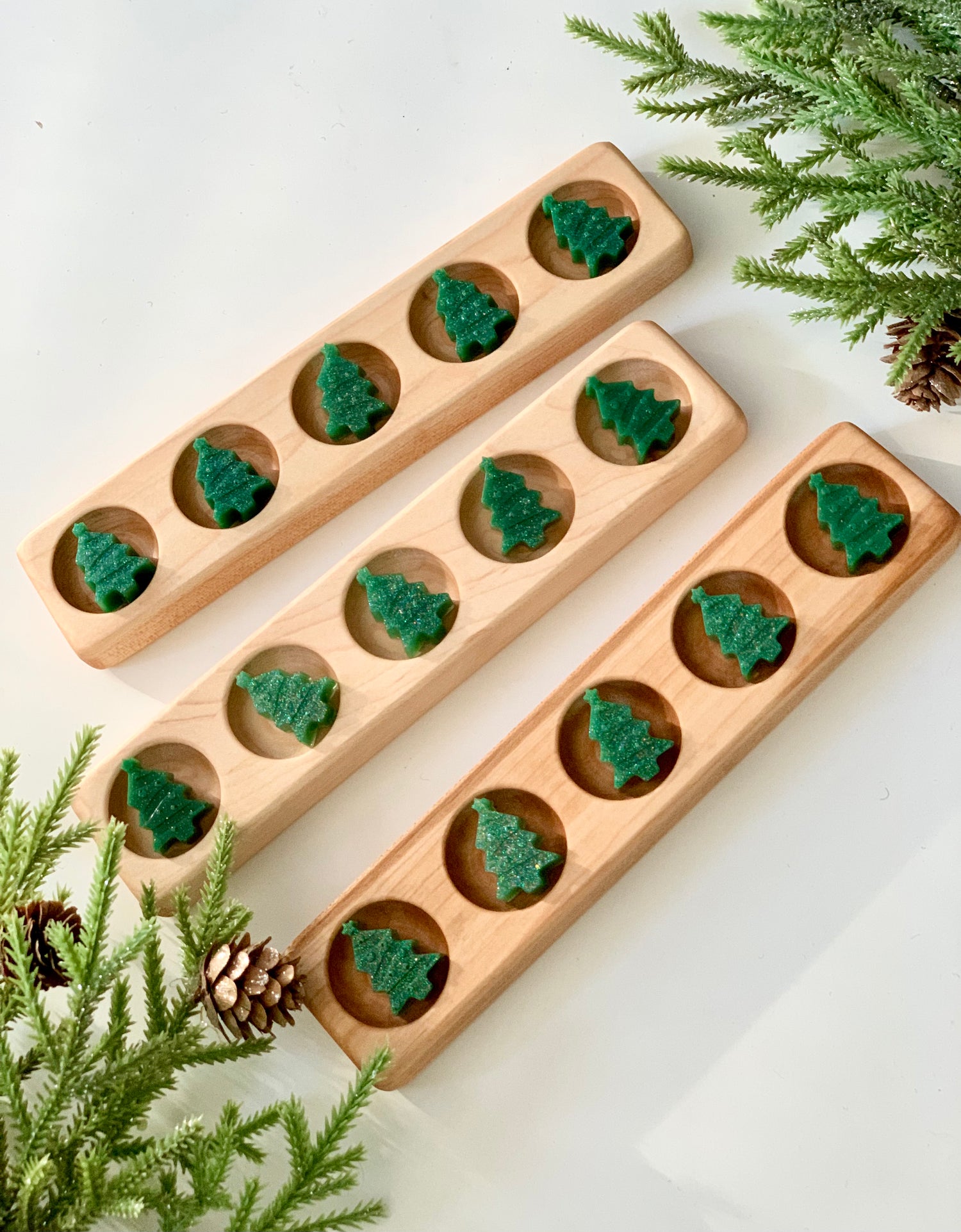 Three wooden mancala boards with green Christmas tree-shaped playing pieces surrounded by pine branches and cones