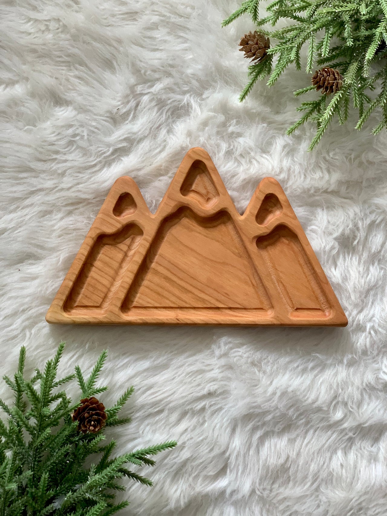Mountain-shaped wooden sensory tray with three carved peaks on white fur background with pine branches and cones