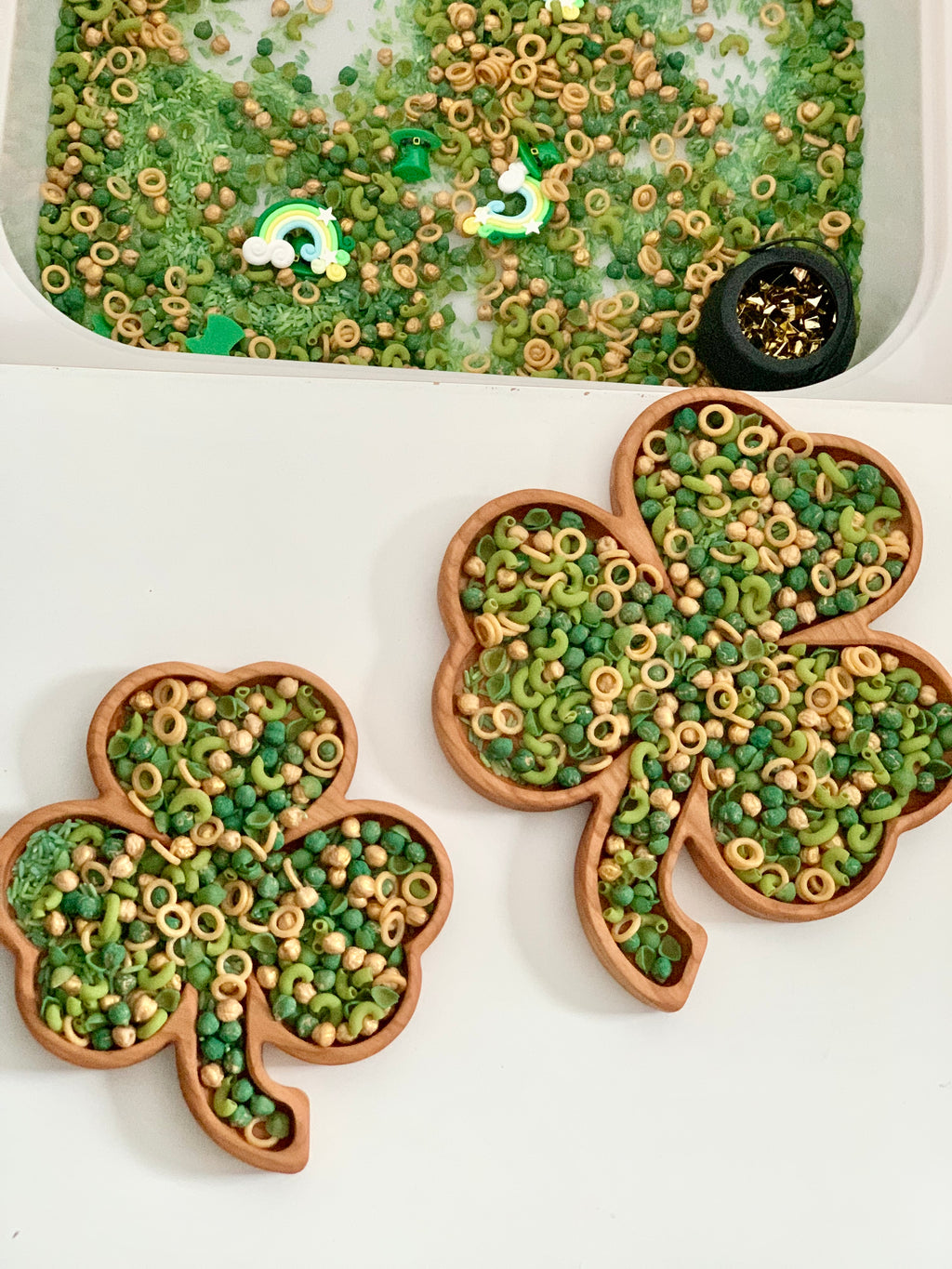 Two wooden shamrock bowls filled with green and beige pasta and beads beside a sensory bin with rainbows and a small black pot