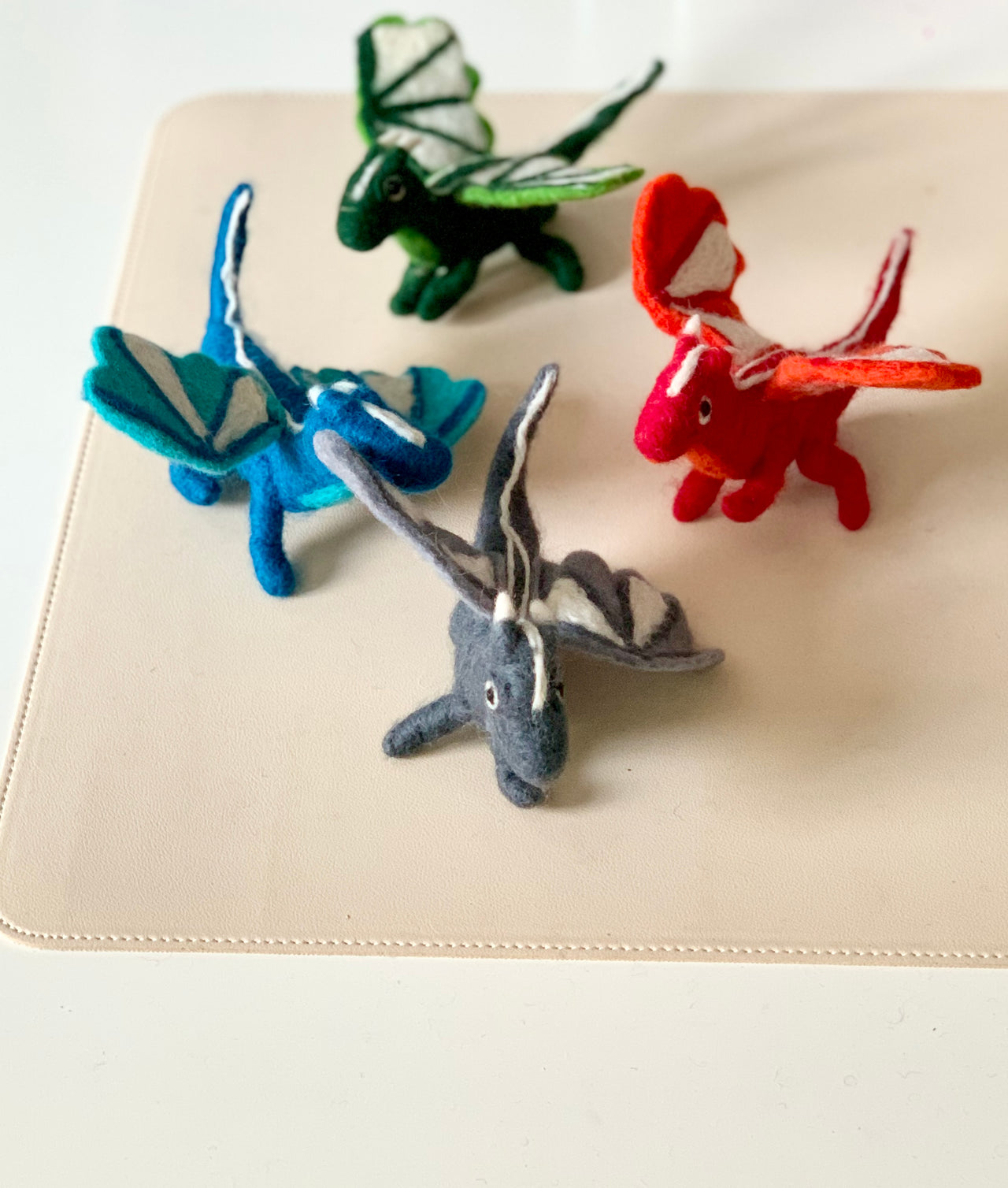 Four colorful handmade felt dragon toys in green, red, blue, and gray with wings on a beige surface