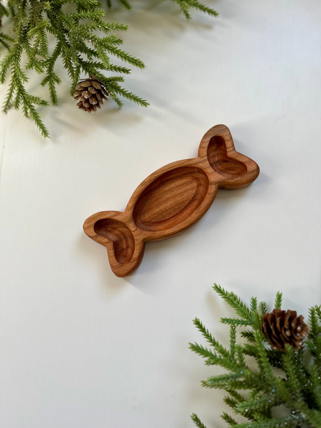 Wooden tray carved in the shape of a wrapped candy surrounded by pine branches and pine cones on white background