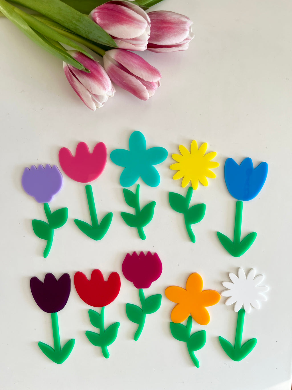 Brightly colored plastic flower shapes with green stems arranged in two rows beside three real pink tulips on white surface