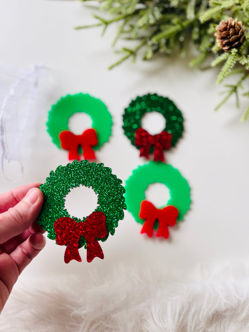 Hand holding small glittery green Christmas wreath with red bow, with similar wreaths and pine branch in background