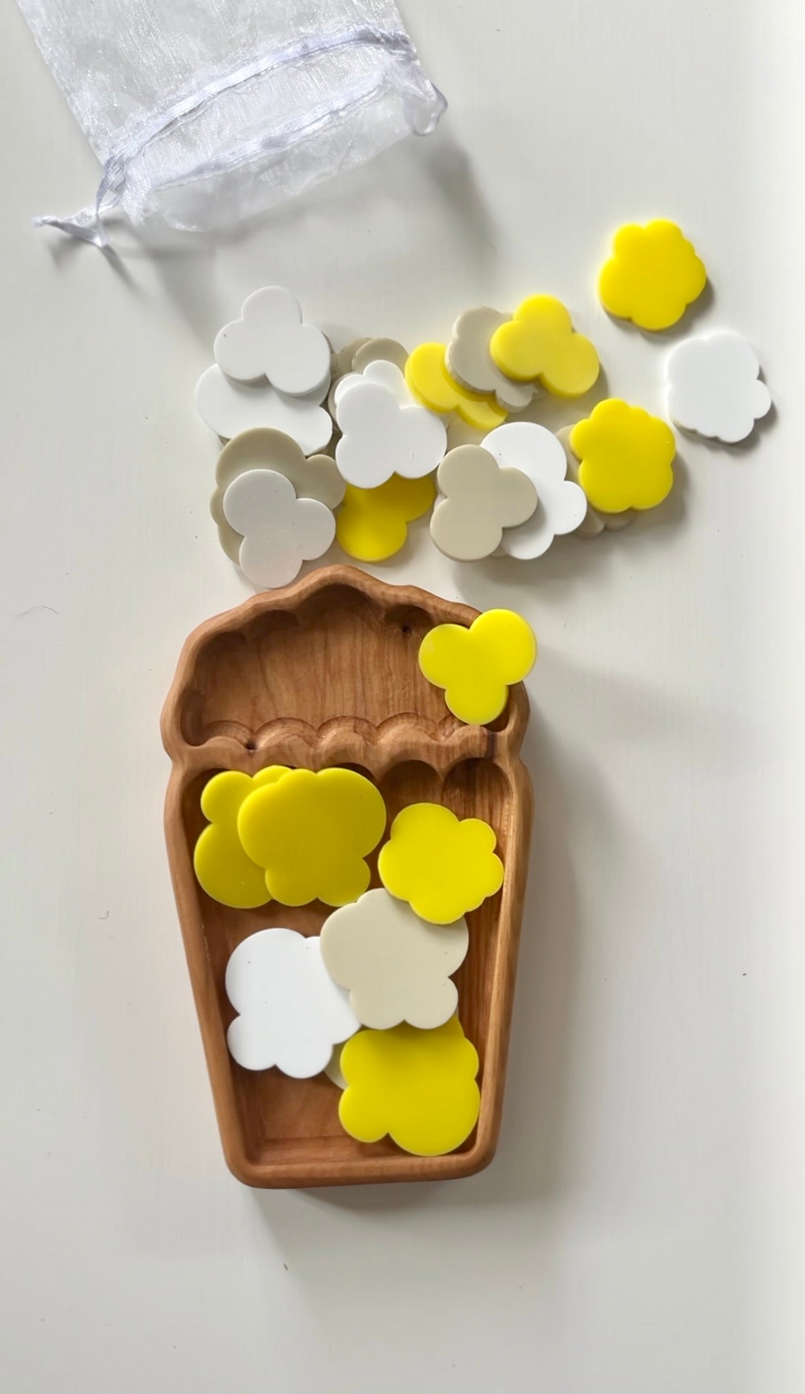 Wooden tray shaped like a cup with yellow, white, and beige clover-shaped tokens spilling from a sheer white bag on white surface