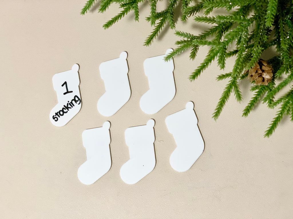 Set of six white dry erasable stocking-shaped cards with one card handwritten number and word on beige surface with greenery nearby