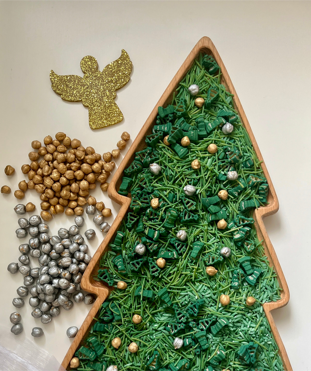 Tree-shaped wooden tray filled with green pasta shapes and gold and silver decorative balls next to a glittery gold angel cutout